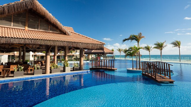 A resort pool area with a thatched-roof structure, palm trees, and a clear view of the ocean. - Aquiraz Manhattan Riviera (Aquiraz)