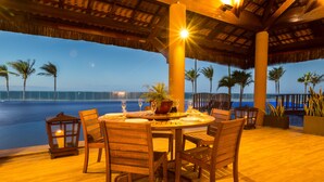 A wooden dining table with chairs, a lantern, and a potted plant on an outdoor deck with a view of the ocean and palm trees. - Aquiraz Manhattan Riviera (Aquiraz)