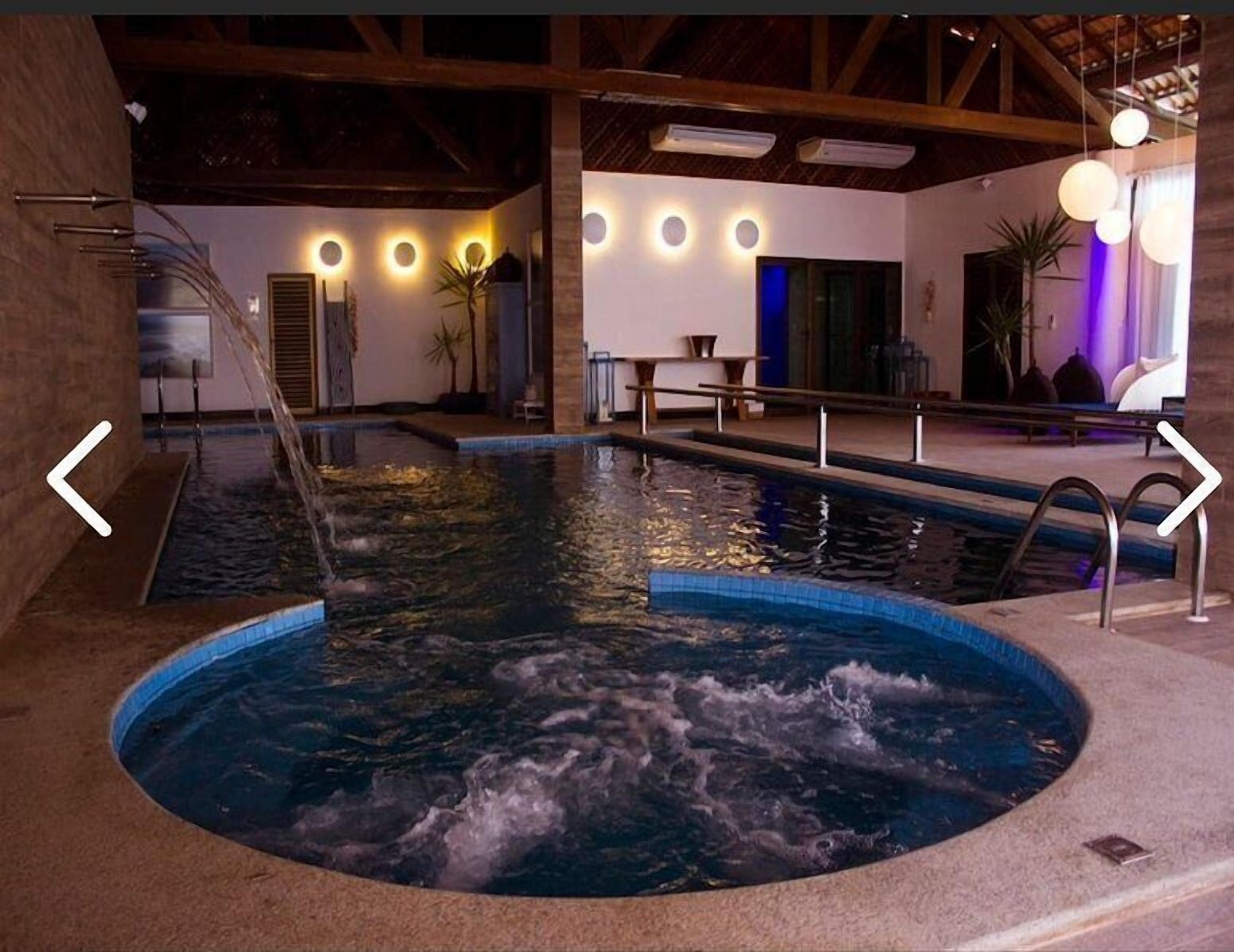 An indoor swimming pool with a hot tub, surrounded by a wooden deck and a lounge area with seating and ambient lighting.