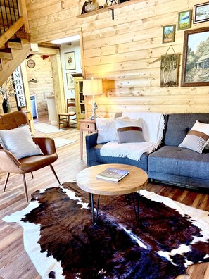 Interior acolhedor de uma cabana de madeira, com um sofá, poltrona e mesa de centro redonda.
