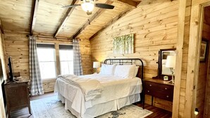 Une chambre dans une cabane en bois, comprenant un lit, une table de chevet, une commode, un ventilateur de plafond et une fenêtre avec des rideaux.