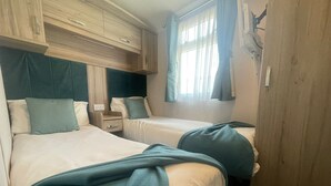 Ein Zimmer mit zwei Betten, jedes mit Kopfkissen und Decke. Es gibt ein hölzernes Kopfteil, ein Fenster mit Vorhängen und ein an der Wand befestigtes Regal.