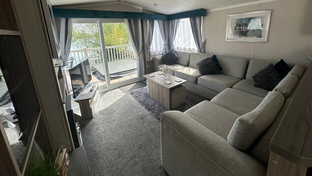 Ein Wohnzimmer mit einer Couch, einem Couchtisch und einem Fernseher. Es gibt einen Balkon mit Geländer und Blick auf Bäume.