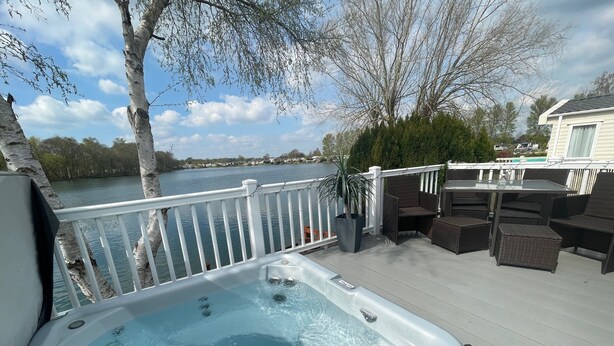Un jacuzzi en una terraza con vistas a un lago, rodeado de muebles de mimbre y una barandilla.