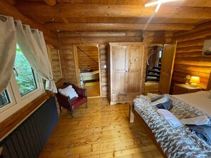 Interior de una cabaña de madera con una cama, una silla roja, un armario de madera y un ventanal con cortinas.