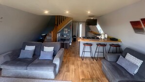 A modern living room with two grey sofas, a wooden coffee table, and a kitchen area with a bar stool and a TV.