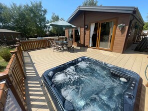 Una vasca idromassaggio su un pontile di legno con tavolo e sedie, un'area patio coperta e un edificio con ampie finestre.