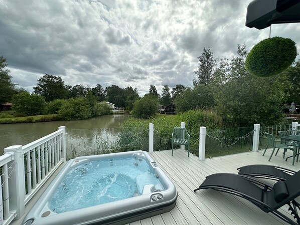 A hot tub on a wooden deck with a river view, surrounded by greenery and a cloudy sky.