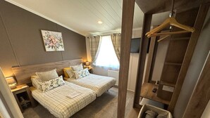 A bedroom with two beds, a bedside table with a lamp, a hanging rack, and a basket with towels. - Luxury lakeside lodge with sunken hot tub prime location on bull rush lake @tattershall lakes (Tattershall)