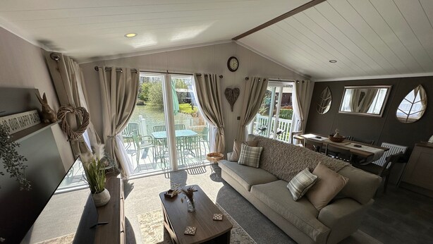 A spacious living area with a sofa, a coffee table, and a dining table. There is a large window with curtains, a clock on the wall, and a view of an outdoor patio. - Luxury lakeside lodge with sunken hot tub prime location on bull rush lake @tattershall lakes (Tattershall)
