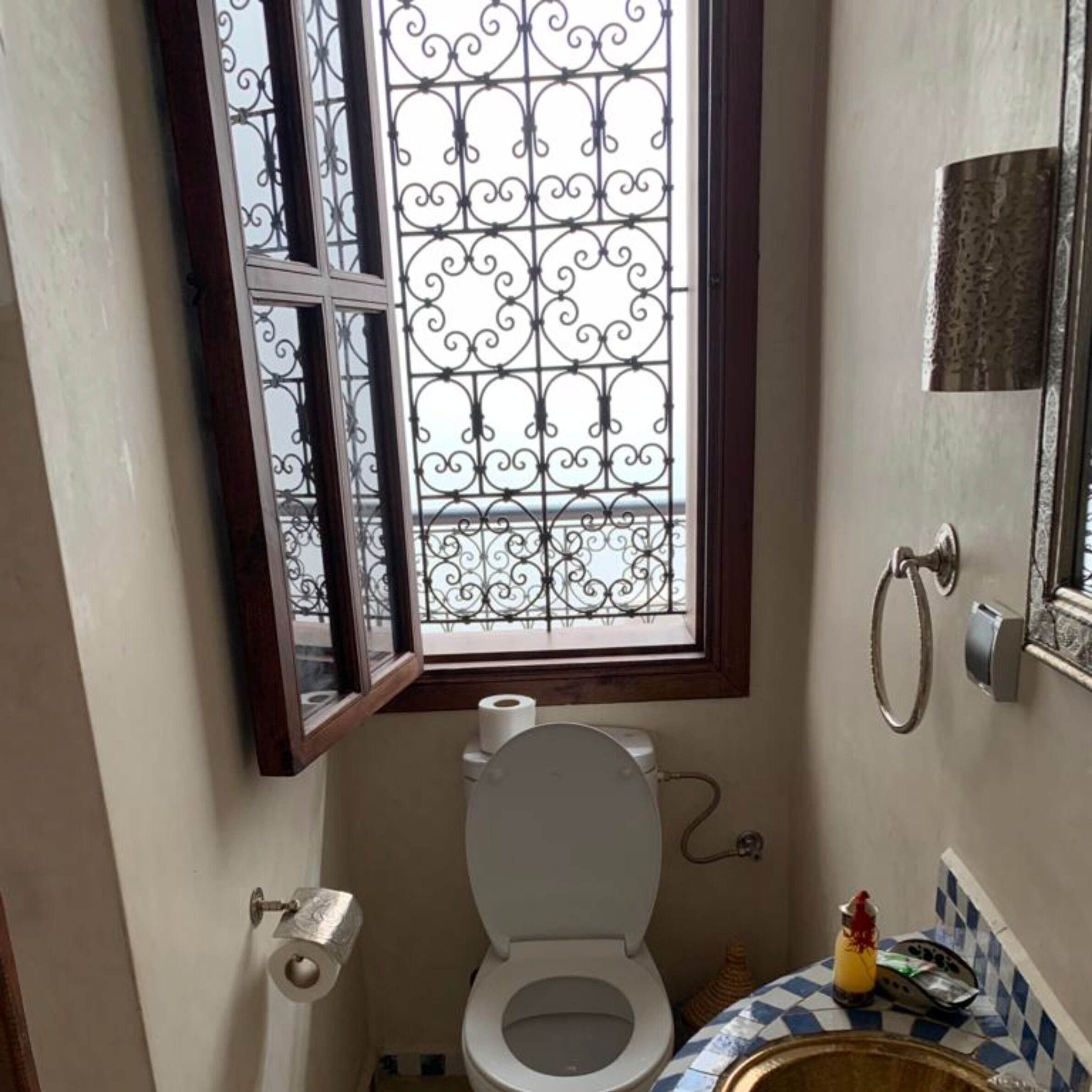 Een kleine badkamer met toilet, een raam met decoratieve ijzeren spijlen, een handdoekhouder en een betegeld wastafelgedeelte.