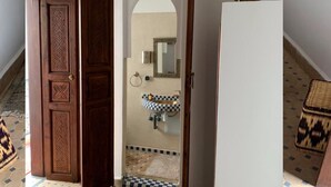 Un couloir doté d’un sol à carreaux, une porte en bois ornée de sculptures complexes, un mur blanc avec un miroir et un petit tapis.