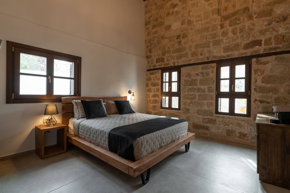Un dormitorio con una cama de madera, dos ventanas, una mesita de noche con una lámpara y una pared de piedra.