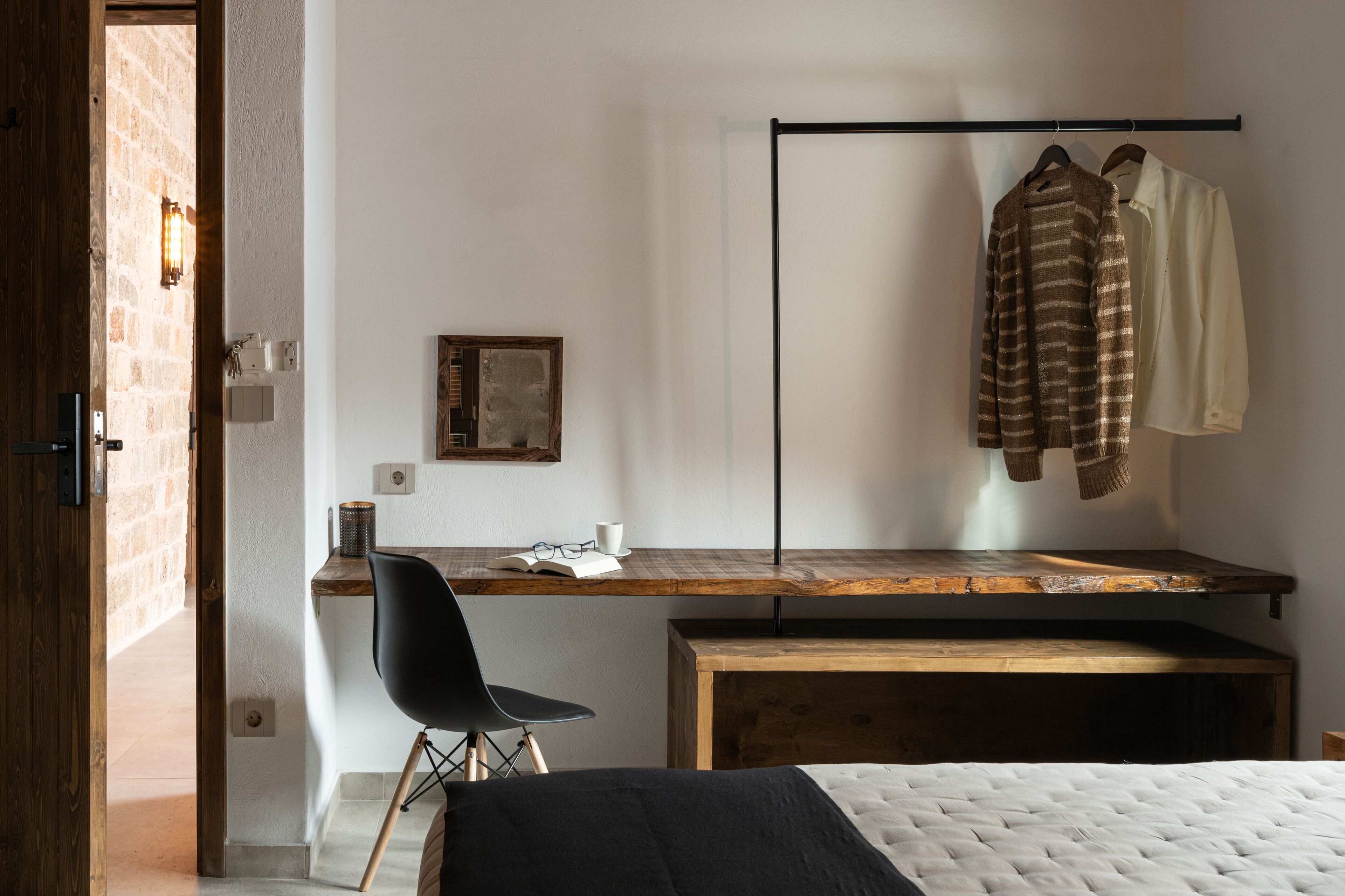 Un dormitorio con un escritorio de madera, una silla negra, una cama con colcha blanca y un perchero con dos camisas.