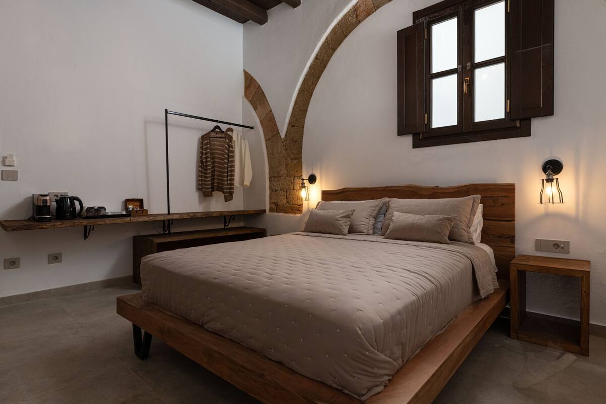 Un dormitorio con cama de madera, una estantería con ropa, una lámpara y una ventana con contraventanas.
