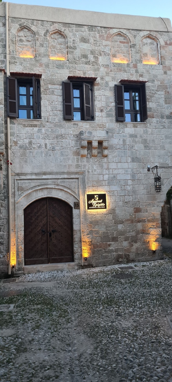 A stone building with a large wooden door, multiple windows with shutters, and a sign indicating the name and number of the establishment. - Ancient Knights Luxury Suites (Rhodes)