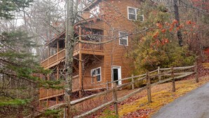 A two-story wooden cabin with a balcony, surrounded by trees and a wooden fence.
