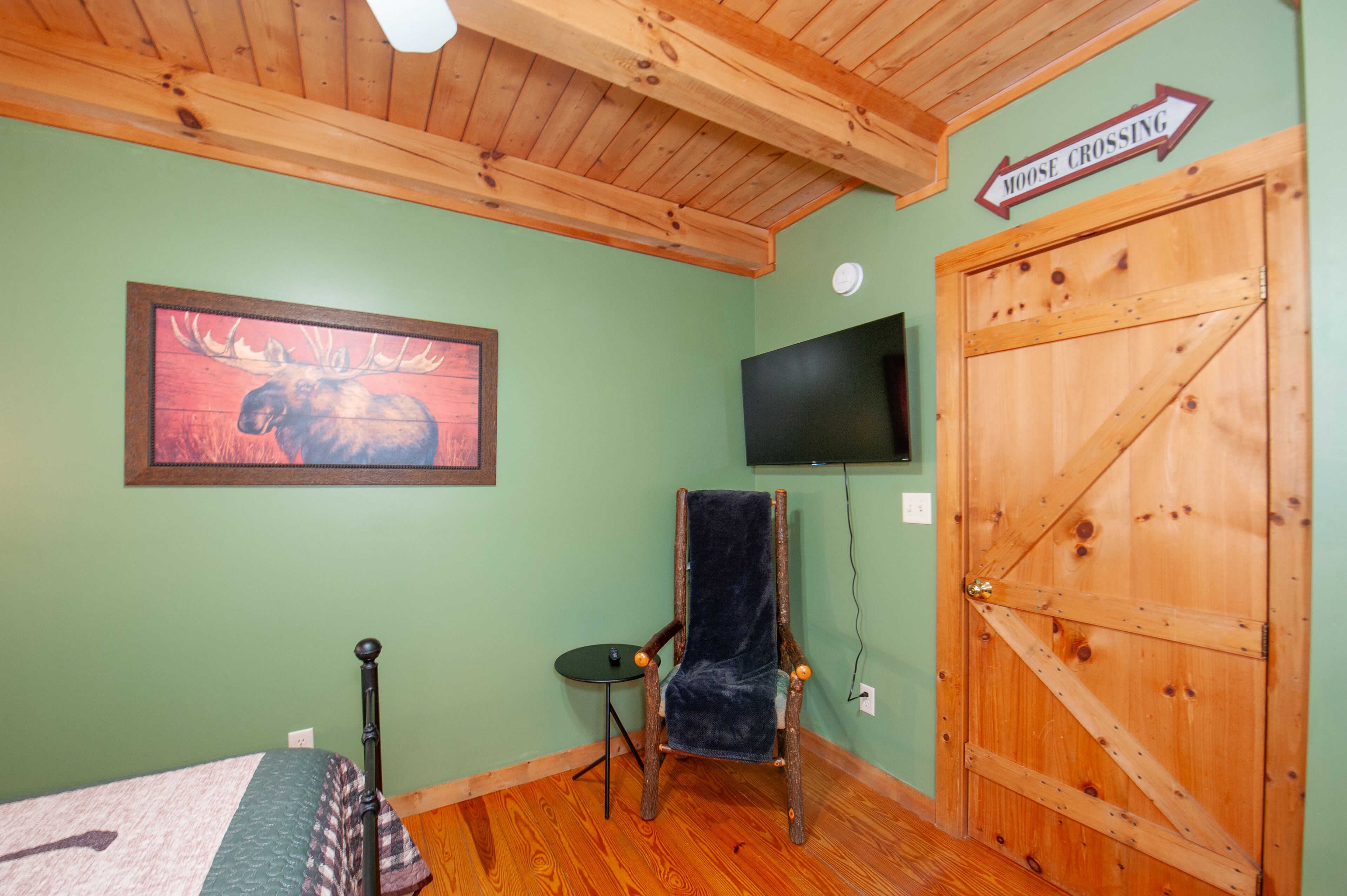 Ein Zimmer mit einem Bett, einem Sessel, einem Fernseher und einer Holztür mit der Aufschrift 'Moose Crossing'.