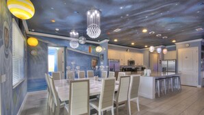 Uma cozinha moderna com armários brancos, uma ilha central com banquetas e uma área de jantar com uma mesa grande e cadeiras.