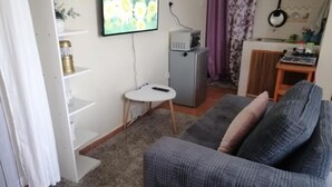 Een woonkamer met een grijze bank, een kleine witte tafel, een televisie aan de muur, en een keukenhoek met een magnetron en een koelkast.