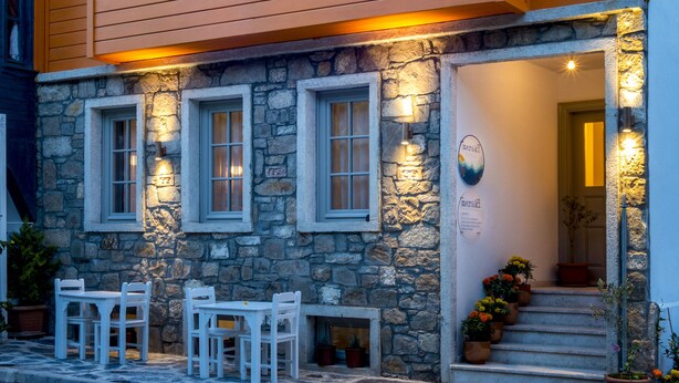 A two-story building with stone and wooden siding, featuring a stone staircase with potted plants, white-framed windows, and outdoor seating. - Meraki Bozcaada (Bozcaada)