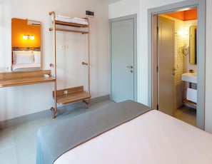 Una camera d'albergo con un letto, una panca di legno, un porta asciugamani, uno specchio, una porta e un bagno visibile attraverso una porta aperta.