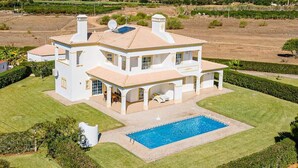 Une grande maison blanche à deux étages, dotée d’une piscine, entourée de pelouses et de jardins bien entretenus.