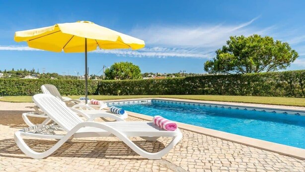 Un espace aménagé au bord de la piscine, avec des chaises longues, un parasol jaune et un ciel d’un bleu limpide.