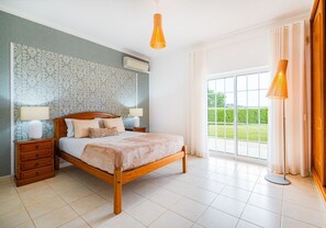 A bedroom with a wooden bed, two bedside tables, a large window with curtains, and a view of greenery outside.