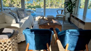 Ein modernes Wohnzimmer mit einem weißen Sofa, blauen Sesseln, einem Couchtisch und Blick auf Palmen und das Meer.