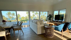 Ein großzügiges Wohnzimmer mit einem weißen Sofa, einem Esstisch mit Stühlen, einem Fernseher und großen Fenstern mit Blick auf Palmen und das Meer.