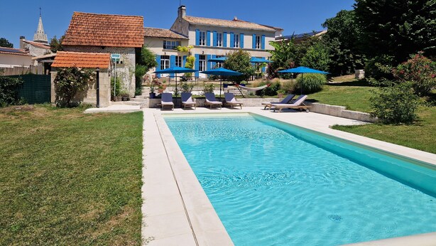 Una piscina con sillas de descanso y sombrillas, rodeada por un césped cuidado y un edificio grande con contraventanas azules.