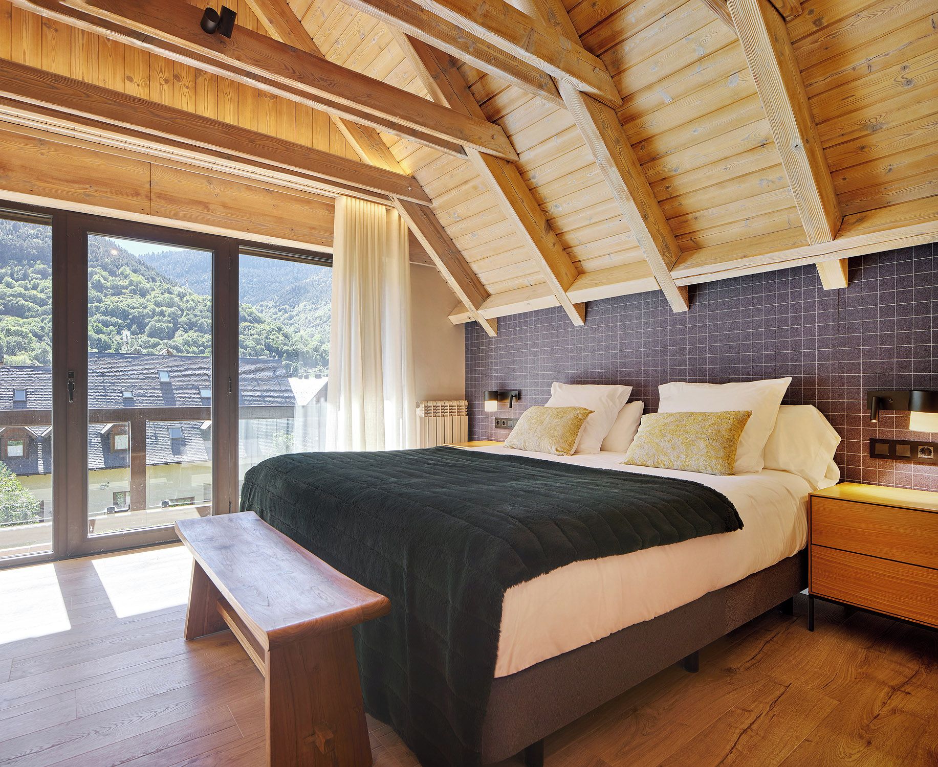 Une chambre avec un grand lit, des poutres apparentes en bois au plafond, une vue sur un balcon et un mur recouvert de carreaux.