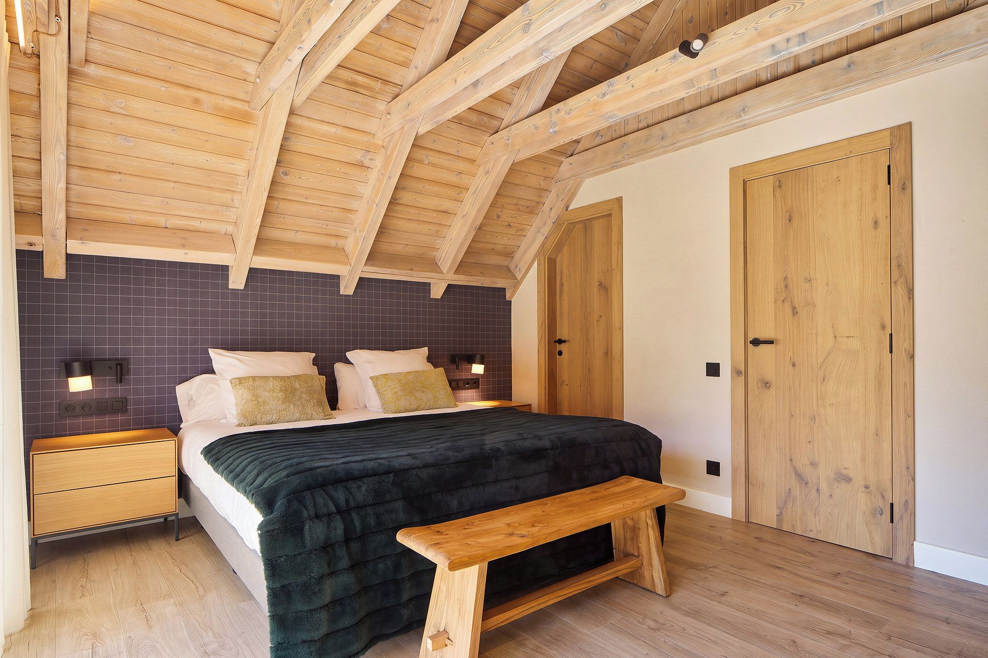 A bedroom with a wooden ceiling, a bed with a dark quilt, two bedside tables with lamps, and a wooden bench.