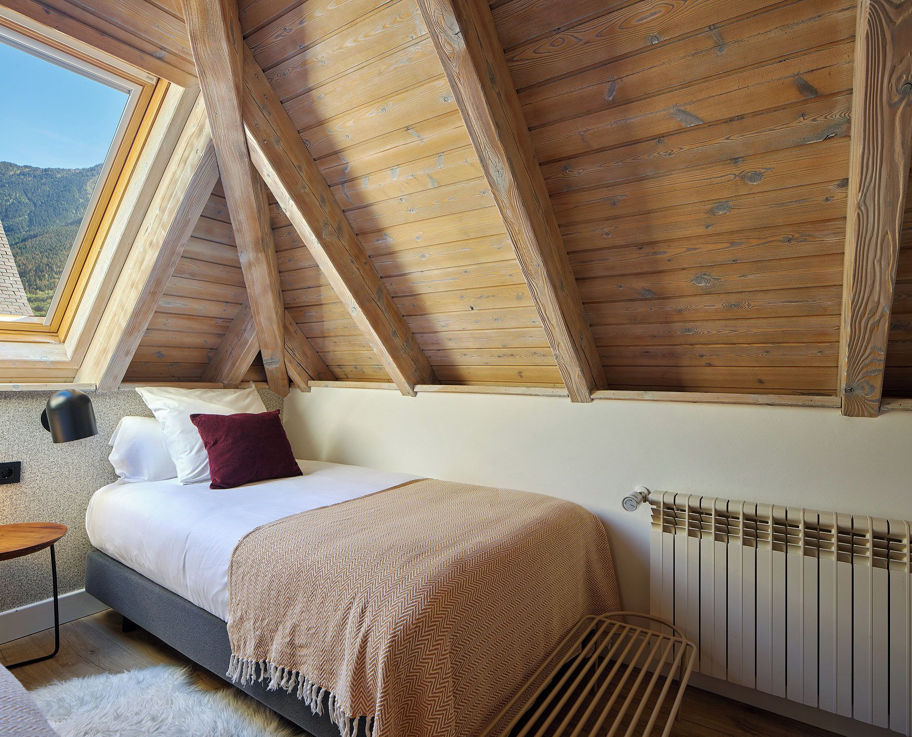 A bedroom with a sloped ceiling, a bed with a white mattress and a beige blanket, a bedside lamp, and a view of the outdoors through a window.