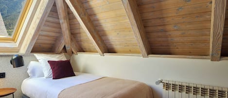 A bedroom with a sloped ceiling, a bed with a white mattress and a beige blanket, a bedside lamp, and a view of the outdoors through a window.