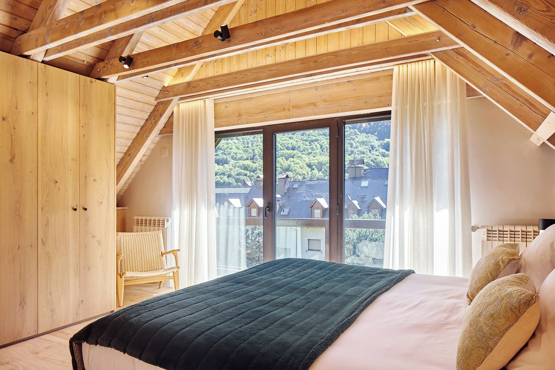 Un dormitorio con un ventanal grande, vigas de madera en el techo, una cama con almohadas y una silla.