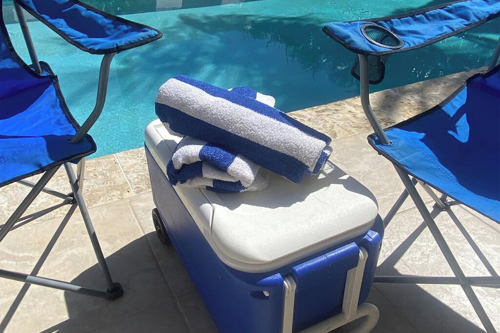 Ein tragbarer Kühler mit einem Handtuch darauf, platziert zwischen zwei blauen Liegestühlen am Pool.