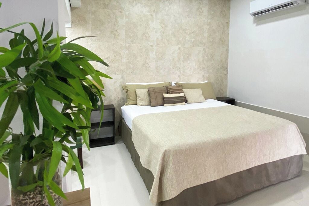 A bedroom with a bed, a bedside table, a nightstand, a potted plant, and an air conditioning unit on the wall.