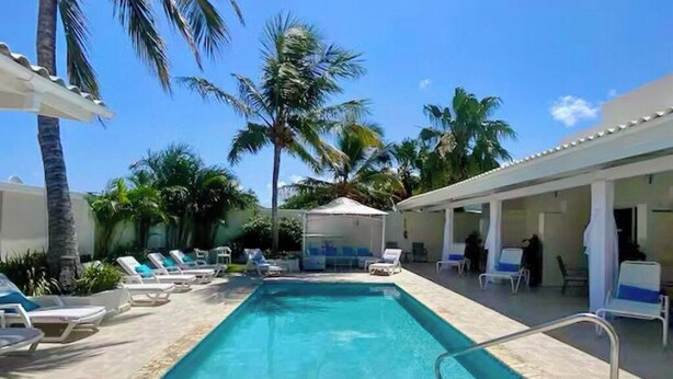 A rectangular swimming pool with a metal ladder, surrounded by lounge chairs and palm trees. - Yoyita Suites Aruba (Noord)