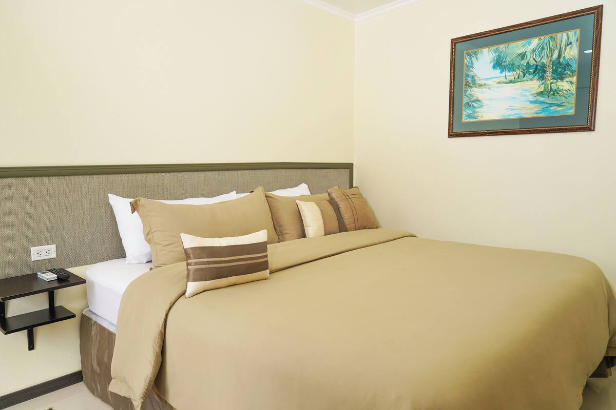 Een netjes opgemaakt bed met een beige deken en kussens, een klein nachtkastje met een afstandsbediening, en een lijst met een schilderij aan de muur.