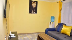 A living room with a sofa, a coffee table, a mounted TV, a water dispenser, and a lion painting on the wall. - Stay.Plus 1 bedroom Imperial Heights (Ruiru)