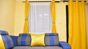 A living room with a sofa, yellow curtains, and a window. - Stay.Plus 1 bedroom Imperial Heights (Ruiru)