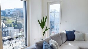 A modern living room with a grey sofa, a large window, and a potted plant. - 2BD high end Apt. 134m2 in Strassen (Strassen)