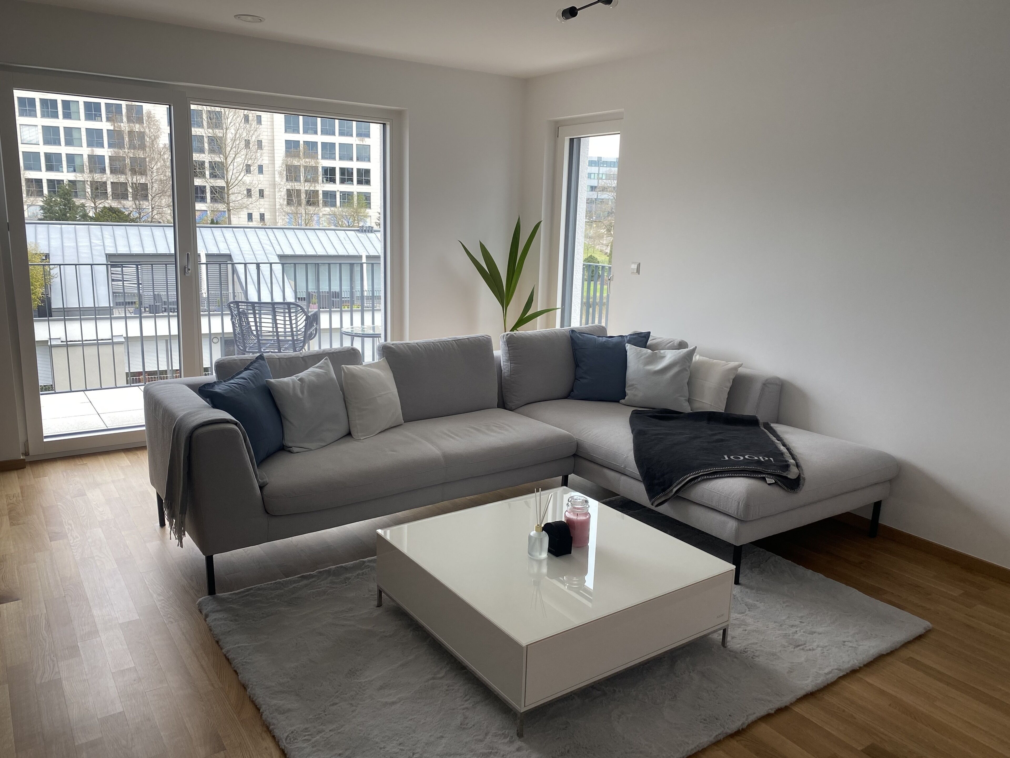 Un salon moderne avec un canapé d’angle gris, une table basse blanche et une grande fenêtre donnant sur un balcon et des immeubles.