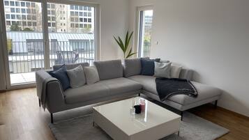 Un salon moderne avec un canapé d’angle gris, une table basse blanche et une grande fenêtre donnant sur un balcon et des immeubles.