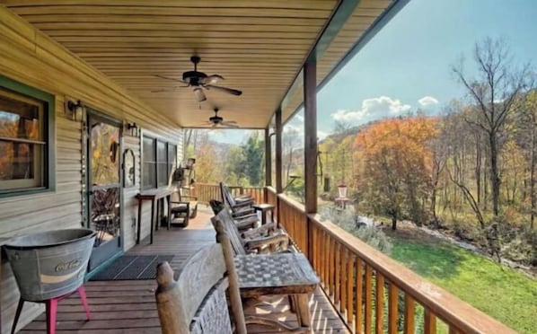 A covered porch with a view of a wooded area, outdoor seating, and a television.