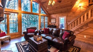 A cozy cabin interior with a leather sofa, wooden walls, large windows, and a staircase. - Private - Creekside - Cabin + Hot Tub/Pool Table & Pet Friendly (Ellijay)