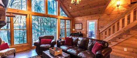 L’intérieur chaleureux d’un chalet avec un canapé en cuir, des murs en bois, de grandes fenêtres et un escalier.