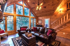 A cozy cabin interior with a leather sofa, wooden walls, large windows, and a staircase. - Private - Creekside - Cabin + Hot Tub/Pool Table & Pet Friendly (Ellijay)